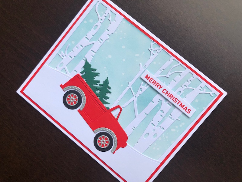 Hand made Christmas card with inked snowfall background, die cut winter trees and truck with Christmas trees.
