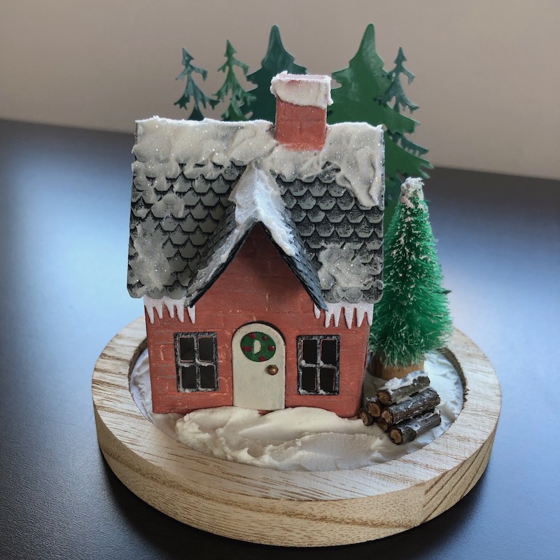 Hand made die cut winter cottage with Christmas trees, log pile and snow.